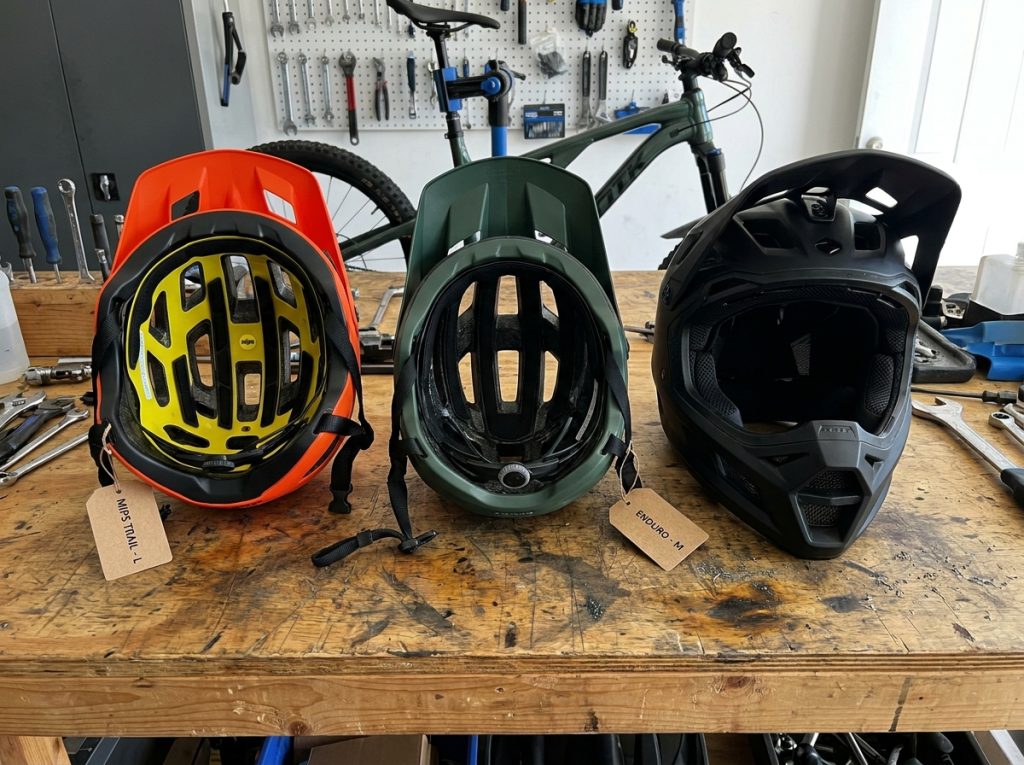Comparaison de trois casques VTT différents posés sur un établi de vélo en bois, trail, enduro et descente